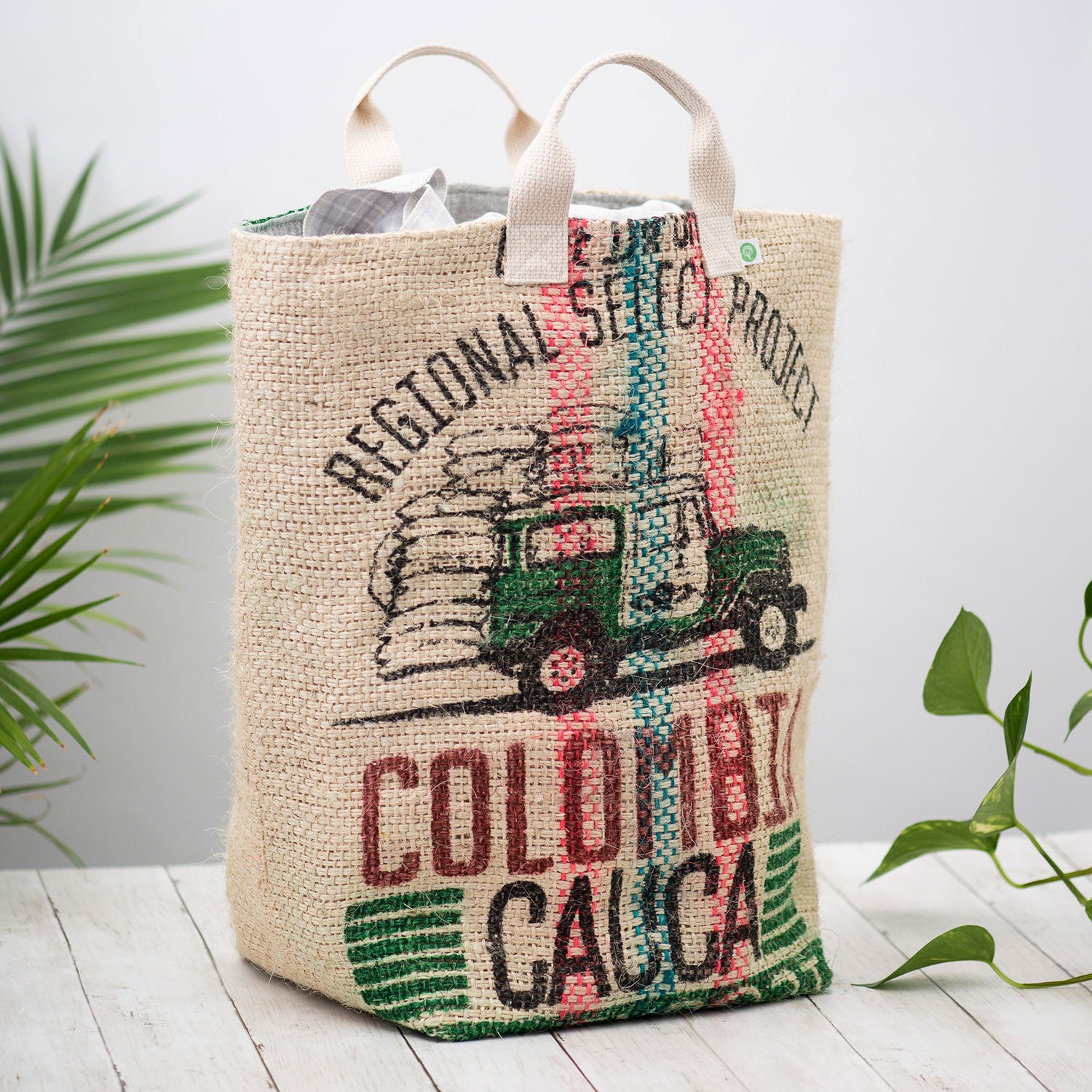 a large burlap coffee sack tote bag stands in a white room among green plants