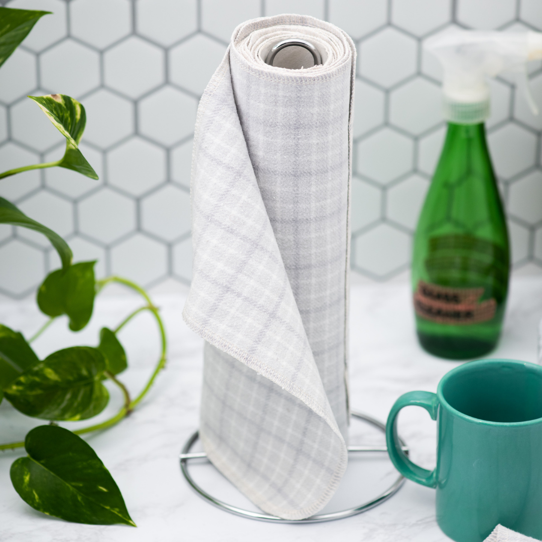 a spool of light grey plaid paperless towels stands upright in the centre with a leafy green plant on the left, and a green glass spray bottle and teal mug on the right